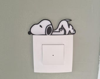 Puede incluir: Una cubierta de interruptor de luz con temática de Snoopy. La cubierta presenta una figura blanca de Snoopy con contornos negros, acostada sobre un interruptor de luz blanco. El fondo es una pared verde claro.