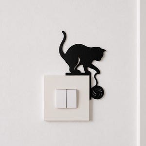 Peut inclure: Une silhouette noire de chat jouant avec une pelote de laine, conçue comme un autocollant mural décoratif qui se fixe sur le bord supérieur d'une plaque d'interrupteur double blanche carrée, avec une queue courbée et une pelote suspendue.
