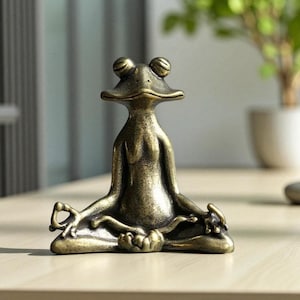 Peut inclure: Figurine de grenouille en bronze dans une pose de yoga assise. La grenouille a de grands yeux ronds et est en position méditative, les jambes croisées et les mains posées sur les genoux. La figurine est en métal et a une finition vieillie.