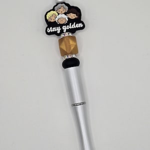 Może przedstawiać: Srebrny długopis z czarną końcówką i ozdobną górą z kreskówkowym wizerunkiem czterech kobiet i napisem "stay golden" czarnymi literami. Długopis ma złoto-czarny wzór z koralików.