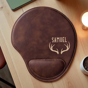 May include: A brown leatherette mouse mat with a wrist rest. The mat features the name "SAMUEL" above a deer antler design. The mat is on a wooden surface.