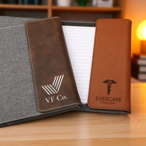 May include: A gray fabric portfolio with two brown leather covers. One cover has the text "VF Co." and a logo, the other has the text "EVERCARE medical clinic" and a medical symbol. The portfolio holds a notepad.