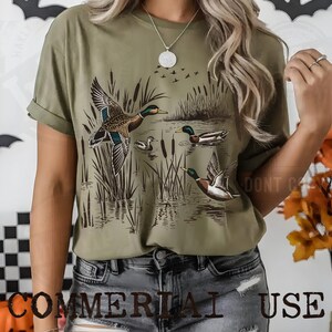 K&ouml;nnte beinhalten: Olivgr&uuml;nes T-Shirt mit einer detaillierten Illustration von Enten in einer Sumpflandschaft. Das Design zeigt Enten im Flug und auf dem Wasser, mit Schilf und einem Vogelschwarm im Hintergrund. Der Text "COMMERCIAL USE" steht unten.