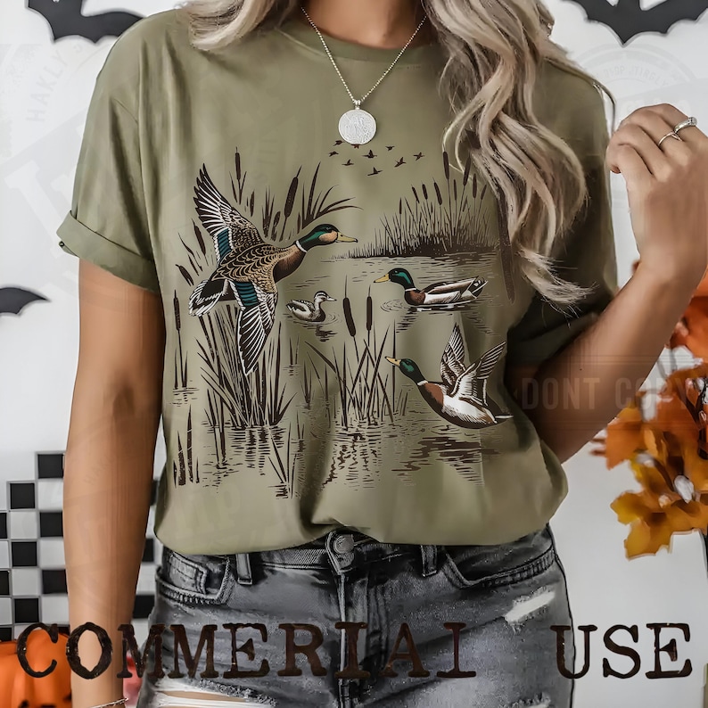 K&ouml;nnte beinhalten: Olivgr&uuml;nes T-Shirt mit einer detaillierten Illustration von Enten in einer Sumpflandschaft. Das Design zeigt Enten im Flug und auf dem Wasser, mit Schilf und einem Vogelschwarm im Hintergrund. Der Text "COMMERCIAL USE" steht unten.