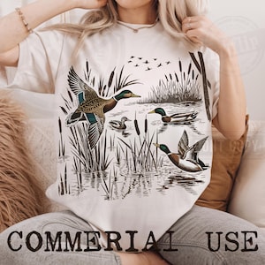 K&ouml;nnte beinhalten: Cremefarbenes T-Shirt mit einer detaillierten Illustration von Enten in einer Sumpflandschaft. Das Design zeigt Enten im Flug und auf dem Wasser, mit Schilf und fliegenden V&ouml;geln. Der Text "COMMERCIAL USE" ist unten aufgedruckt.