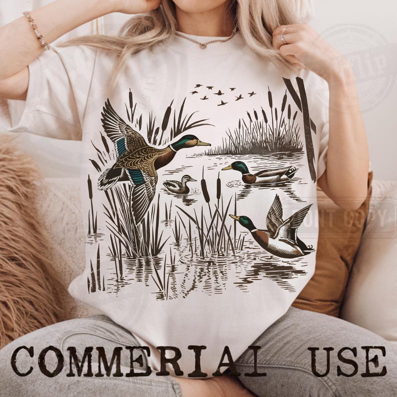 K&ouml;nnte beinhalten: Cremefarbenes T-Shirt mit einer detaillierten Illustration von Enten in einer Sumpflandschaft. Das Design zeigt Enten im Flug und auf dem Wasser, mit Schilf und fliegenden V&ouml;geln. Der Text "COMMERCIAL USE" ist unten aufgedruckt.