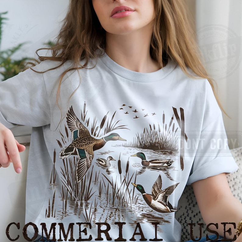 K&ouml;nnte beinhalten: Hellblaues T-Shirt mit einer detaillierten Illustration von Enten in einer Sumpflandschaft. Das Design zeigt Enten im Flug und auf dem Wasser, mit Schilf und einem Vogelschwarm im Hintergrund. Die Worte "COMMERCIAL USE" sind unten aufgedruckt.
