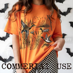 K&ouml;nnte beinhalten: Leuchtend orangefarbenes T-Shirt mit einer detaillierten Illustration von Enten in einem Sumpf. Die Grafik zeigt Enten im Flug und auf dem Wasser, mit Schilf und fliegenden V&ouml;geln. Der Text "COMMERIA HOUSE" befindet sich unten.