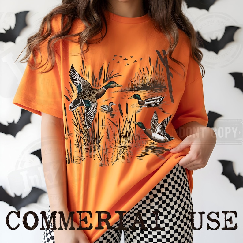 K&ouml;nnte beinhalten: Leuchtend orangefarbenes T-Shirt mit einer detaillierten Illustration von Enten in einem Sumpf. Die Grafik zeigt Enten im Flug und auf dem Wasser, mit Schilf und fliegenden V&ouml;geln. Der Text "COMMERIA HOUSE" befindet sich unten.