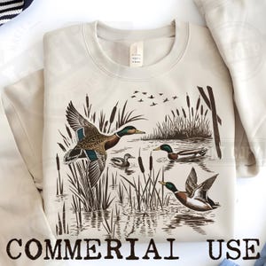 K&ouml;nnte beinhalten: Beiges Sweatshirt mit einer detaillierten Illustration von Enten in einer Sumpflandschaft. Das Design zeigt Enten im Flug und auf dem Wasser, mit Schilf und fliegenden V&ouml;geln. Die Worte "COMMERCIAL USE" sind unten aufgedruckt.