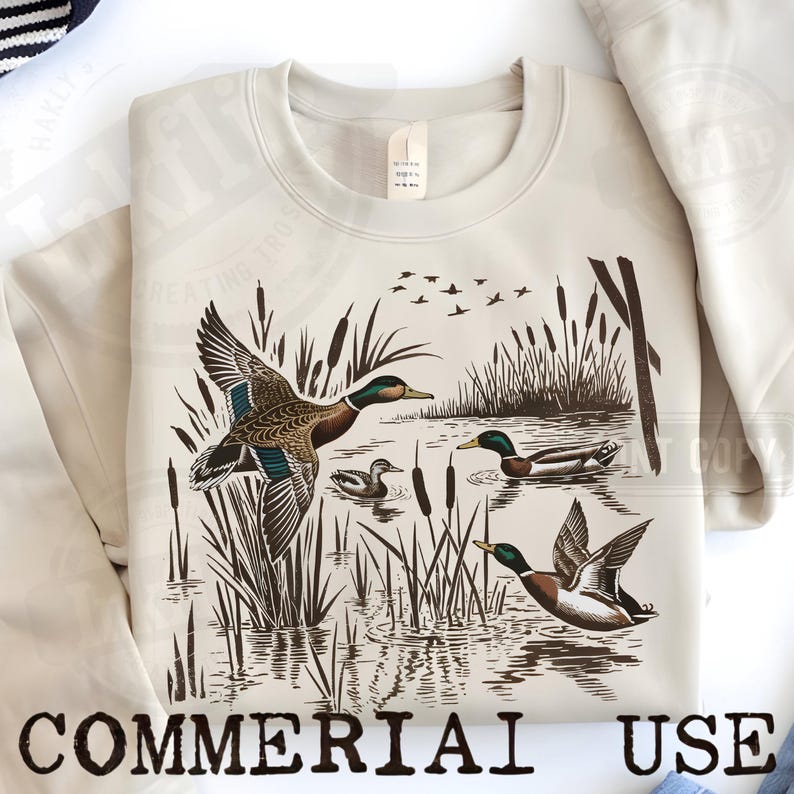 K&ouml;nnte beinhalten: Beiges Sweatshirt mit einer detaillierten Illustration von Enten in einer Sumpflandschaft. Das Design zeigt Enten im Flug und auf dem Wasser, mit Schilf und fliegenden V&ouml;geln. Die Worte "COMMERCIAL USE" sind unten aufgedruckt.