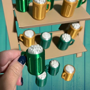 May include: A collection of miniature beer mugs in green and gold, some filled with white foam. The mugs are arranged on a tiered display shelf, with a hand holding one of the green mugs. The background is a blue and green surface.