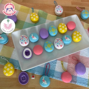May include: Easter-themed macarons and egg-shaped ornaments in various pastel colors. Some macarons feature floral or bunny designs. The ornaments have keychains attached. The image includes the text "Marie's Button Authorized Seller" and "Tidy's 3D Prints".