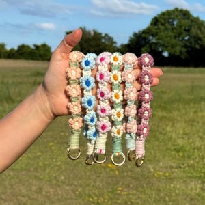 May include: A selection of daisy-patterned keyrings in pastel shades of pink, blue, and peach. Each keyring has a gold-coloured ring. The keyrings are made of a woven material and are shown against a green field and blue sky.