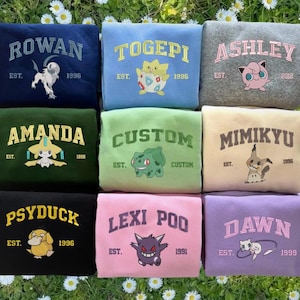 May include: A collection of nine sweatshirts in various colors, each featuring a name or word in a collegiate font, along with a graphic of a cartoon character. The sweatshirts include names like "Rowan," "Togepi," and "Ashley," with the year of establishment printed below.
