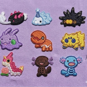 May include: Nine embroidered patches featuring various cartoon creatures in vibrant colors. The patches are on a purple fabric background. The creatures include a black and pink spiky design, a blue cloud with a party hat, and a purple rabbit-like figure.