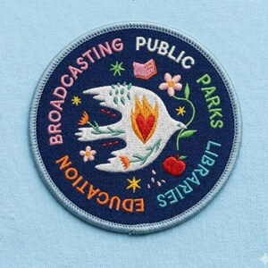 May include: Embroidered patch with a navy blue border and a light blue background. The patch features a white dove with a red heart and orange flames. The words "Broadcasting Public Parks Libraries Education" are embroidered around the edge.