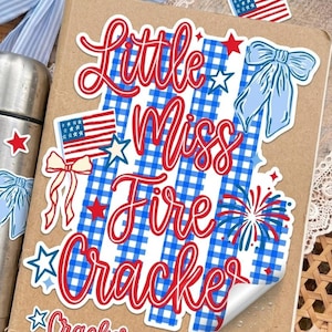 May include: A sticker featuring the red text Little Miss Fire Cracker over a blue and white gingham background. The design includes a small American flag, light blue bows, red and blue stars, and a firework burst, all on a tan notebook cover.