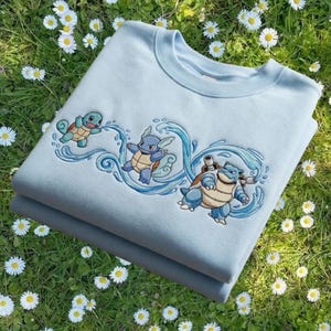 May include: A light blue sweatshirt featuring embroidered images of three cartoon turtle-like creatures with water elements. The sweatshirt is folded and resting on a bed of green grass and white flowers.
