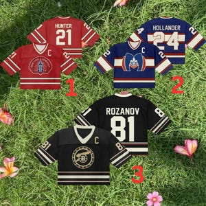 May include: Four short-sleeved jerseys are displayed on grass. The jerseys are red, blue, and black, with white and gold accents. Each jersey features a different design, including numbers, names, and team logos. The jerseys are arranged in a visually appealing composition.