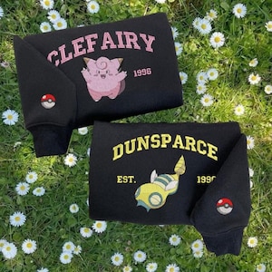 May include: Two black sweatshirts with embroidered designs. One features "CLEFAIRY 1996" with a pink Clefairy character and a Pokéball patch. The other displays "DUNSPARCE EST. 1990" with a yellow and green Dunsparce character and a Pokéball patch.