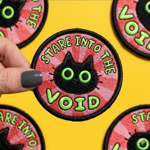 May include: Embroidered patch with a black cat design and the text "STARE INTO THE VOID" in green. The patch has a red and pink radial background and a black border. The cat has green eyes.