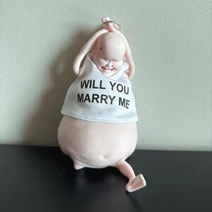 May include: A novelty figurine of a smiling baby wearing a white shirt with the text "WILL YOU MARRY ME". The figurine is light pink and has a ring attached to the top of its head. The baby is in a playful pose.