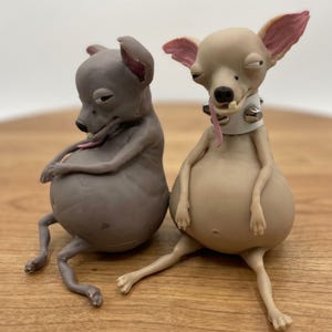 May include: Two stylized dog figurines with large, rounded bellies and thin limbs. One figure is dark gray, and the other is light tan with a white spiked collar. Both have expressive faces with half-closed eyes, protruding tongues, and single visible teeth.