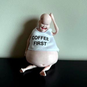 May include: A whimsical figurine of a bald, smiling figure with a large belly. The figure wears a white tank top with the words "COFFEE FIRST" in black. The figure is seated with one arm raised.
