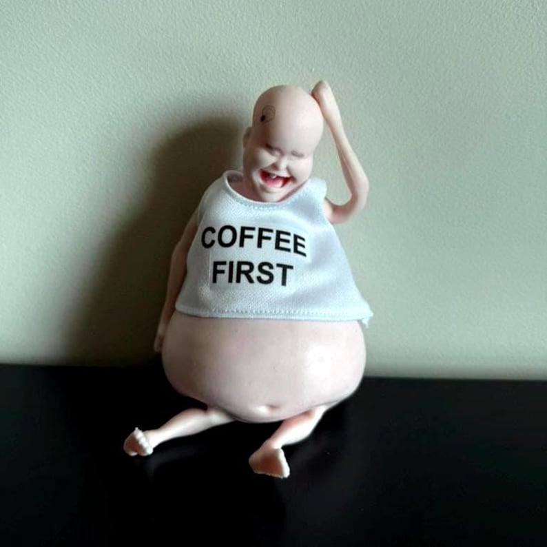 May include: A whimsical figurine of a bald, smiling figure with a large belly. The figure wears a white tank top with the words "COFFEE FIRST" in black. The figure is seated with one arm raised.