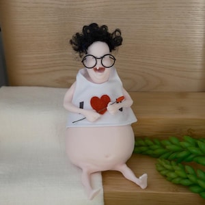 Puede incluir: Una figura caprichosa de una persona con cuerpo redondo, gafas y una camisa blanca con un corazón rojo. La figura tiene cabello negro rizado y sostiene un pequeño objeto rojo. La figura está sobre una superficie de madera.