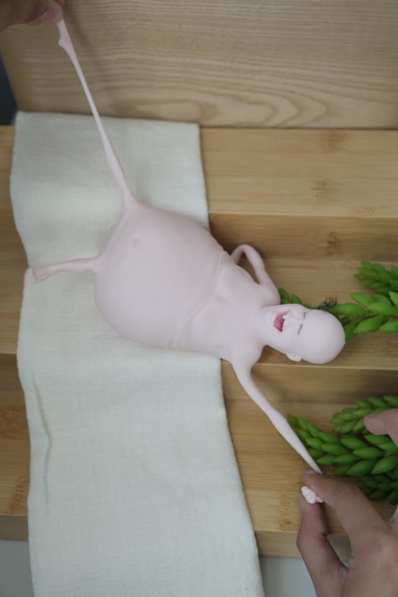 May include: A light pink, abstract sculpture of a human-like figure with elongated limbs and a large torso. The figure is lying on a white cloth, with a wooden surface in the background. Green plant sprigs are on the right.