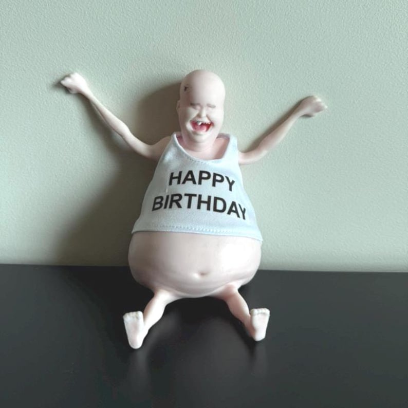 May include: A whimsical figurine of a smiling figure with arms outstretched. The figure wears a white tank top with the words "HAPPY BIRTHDAY" in black. The figurine is light pink and has white feet.