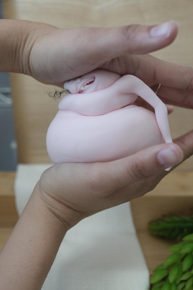 May include: A light pink, abstract sculpture with a rounded body and small arms. The sculpture is held in two hands, with the fingers gently cupping the form. The sculpture has a small face with a slight smile.