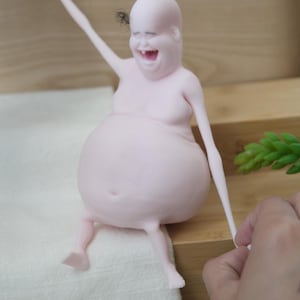 May include: A light pink, smiling figurine with a large belly and thin limbs. The figurine is seated and has one arm raised. A small insect is near the figurine's head. The figurine is being held by a person.