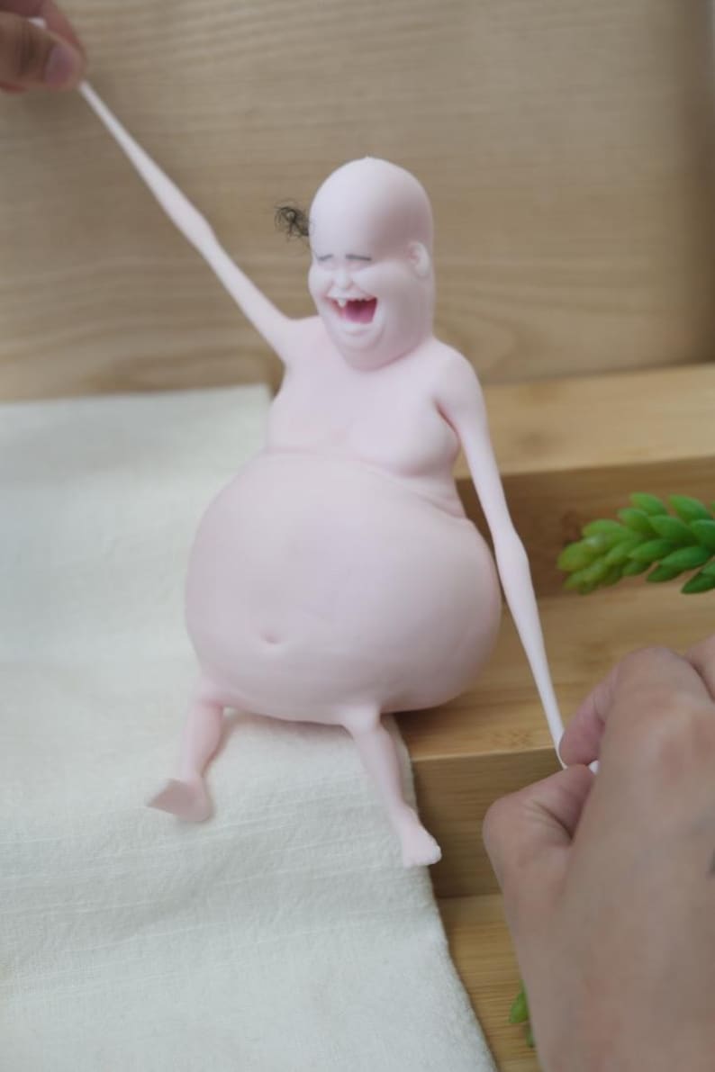 May include: A light pink, smiling figurine with a large belly and thin limbs. The figurine is seated and has one arm raised. A small insect is near the figurine's head. The figurine is being held by a person.