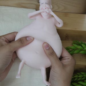 May include: A light pink, cartoonish figurine of a person with a large belly and small legs. The figure's arms are raised to its face, and it has a small tuft of hair. The figurine is being held by two hands.