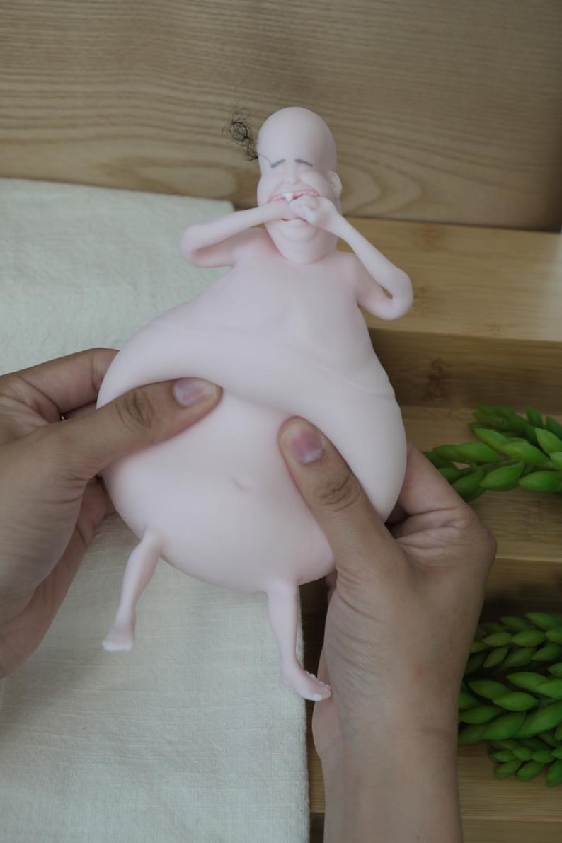 May include: A light pink, cartoonish figurine of a person with a large belly and small legs. The figure's arms are raised to its face, and it has a small tuft of hair. The figurine is being held by two hands.