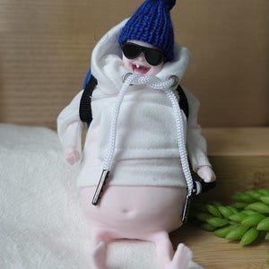 May include: A whimsical doll figure wearing a blue knit beanie, black sunglasses, and a white hooded sweatshirt. The doll has a backpack and is sitting with its legs extended. The figure is on a white surface with a wooden background.