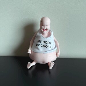 May include: A whimsical figurine of a smiling, bald figure with a large belly and small legs. The figure wears a white tank top with the text "MY BODY MY CHOICE" in black letters. The figure is sitting on a black surface.