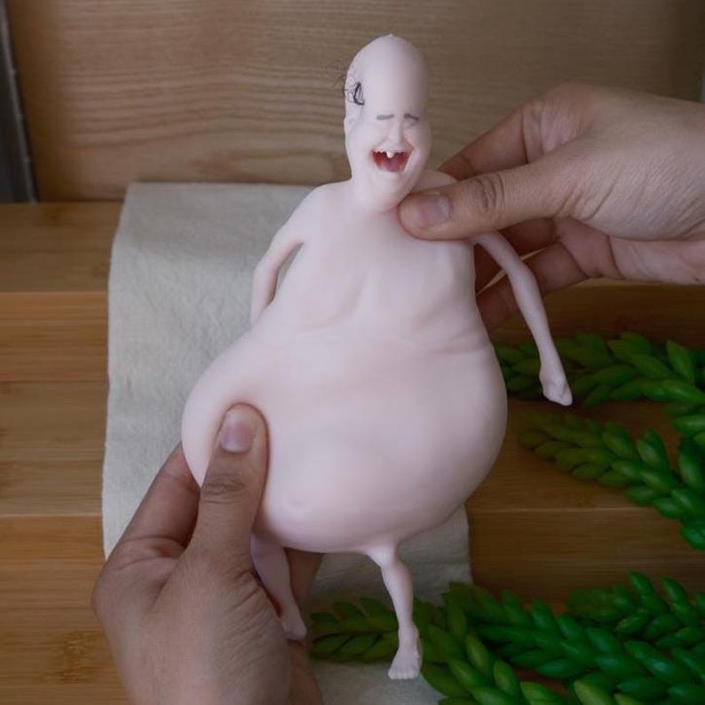 May include: A pale pink, grotesque figurine with a bald head, a wide, toothy grin, and a large, bulbous body. The figure has thin arms and legs and is held by two hands. The background includes a white cloth and green decorative elements.
