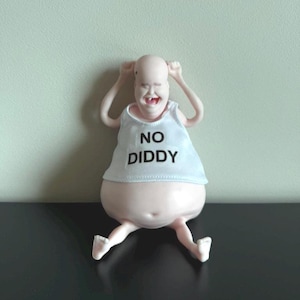 May include: A whimsical figurine of a bald, smiling figure with arms raised. The figure wears a white tank top with the text "NO DIDDY" in black. The figure has a large belly and is seated with legs extended.