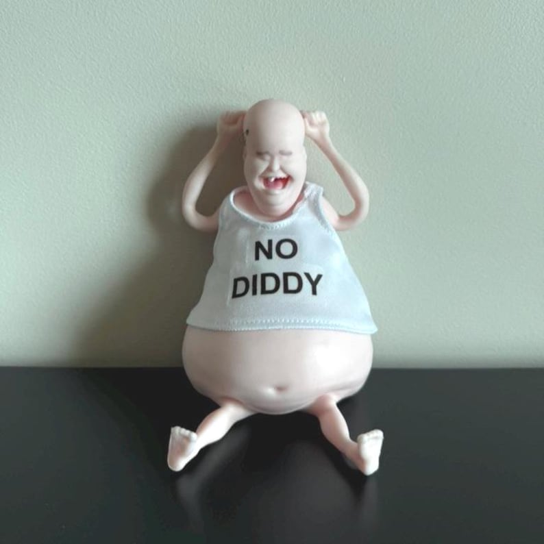 May include: A whimsical figurine of a bald, smiling figure with arms raised. The figure wears a white tank top with the text "NO DIDDY" in black. The figure has a large belly and is seated with legs extended.