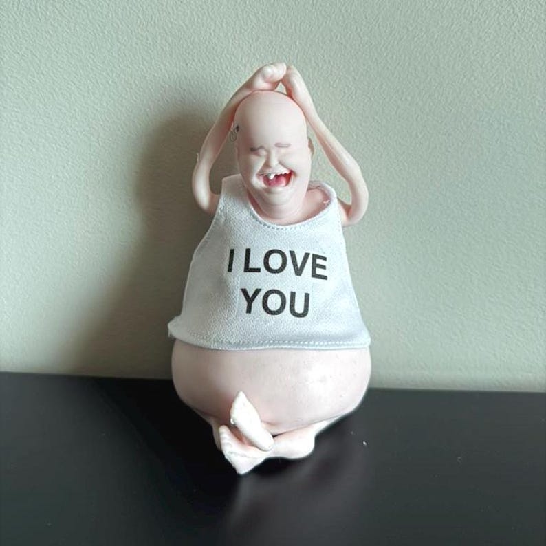 May include: A whimsical figurine of a bald person in a yoga pose, wearing a white tank top with the words "I LOVE YOU" in black. The figure is light pink and has a smiling expression.