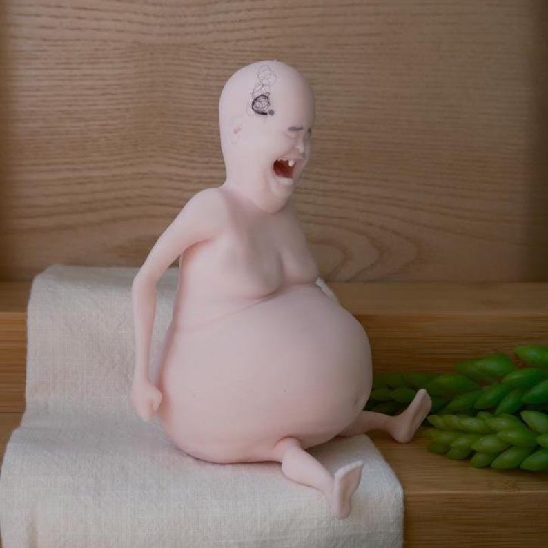May include: A pink figurine of a person with a large belly and open mouth, seated on a white cloth. The figurine has a small drawing on its head and is positioned against a wooden background.