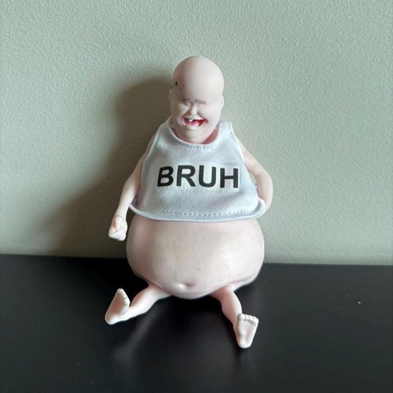 May include: A novelty figurine of a bald, smiling figure with a large belly and small limbs. The figure wears a white bib with the word "BRUH" printed in black. The figurine is sitting on a black surface.