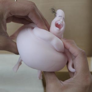 May include: A light pink sculpture of a screaming face with a large open mouth and small black hair. The sculpture is held by two hands, with a gold ring visible on one finger. The sculpture has a rounded body.