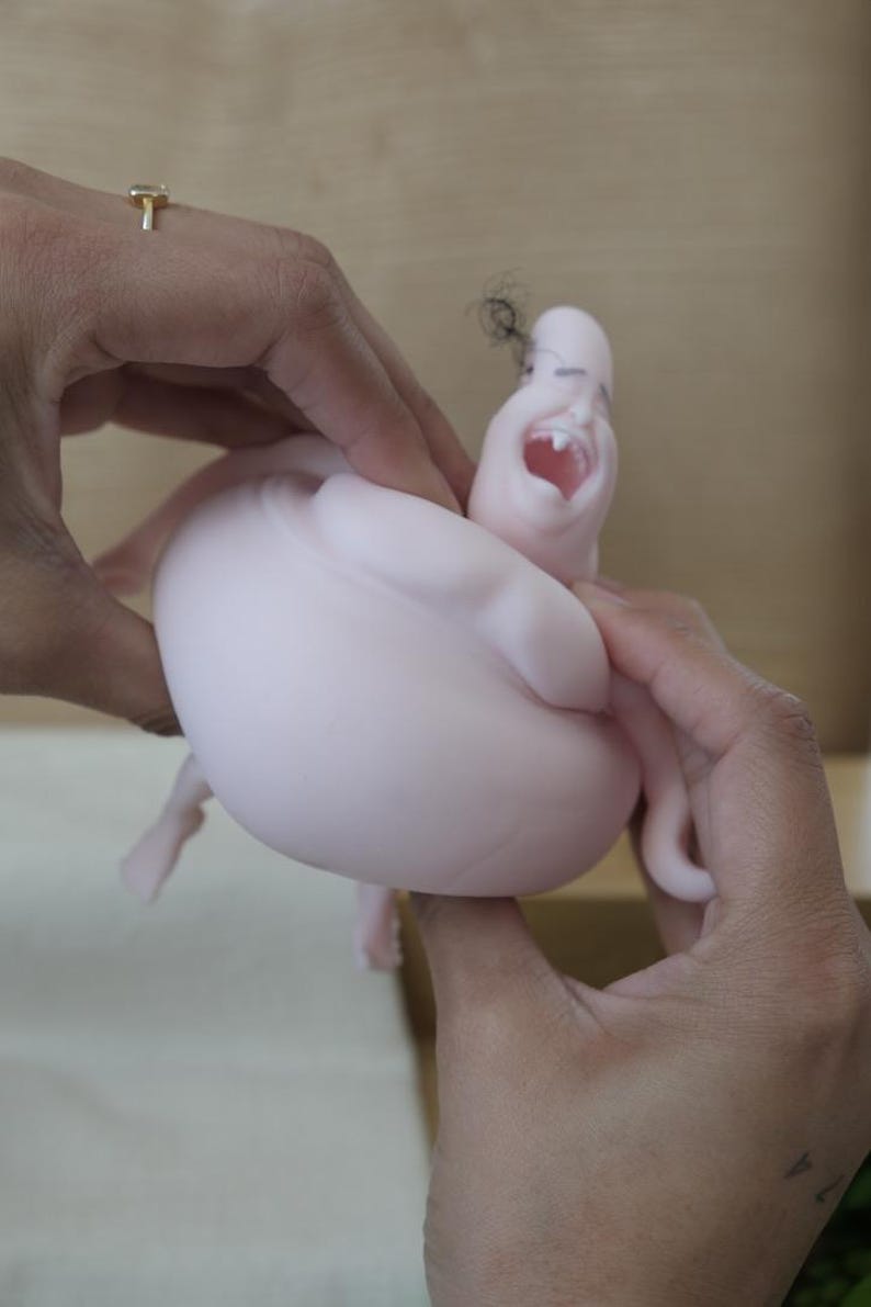 May include: A light pink sculpture of a screaming face with a large open mouth and small black hair. The sculpture is held by two hands, with a gold ring visible on one finger. The sculpture has a rounded body.