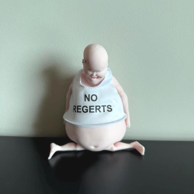 May include: A whimsical figurine of a baby-like figure with a bald head and a smiling expression. The figure wears a white tank top with the words "NO REGERTS" in black. The figure is posed with its legs spread.