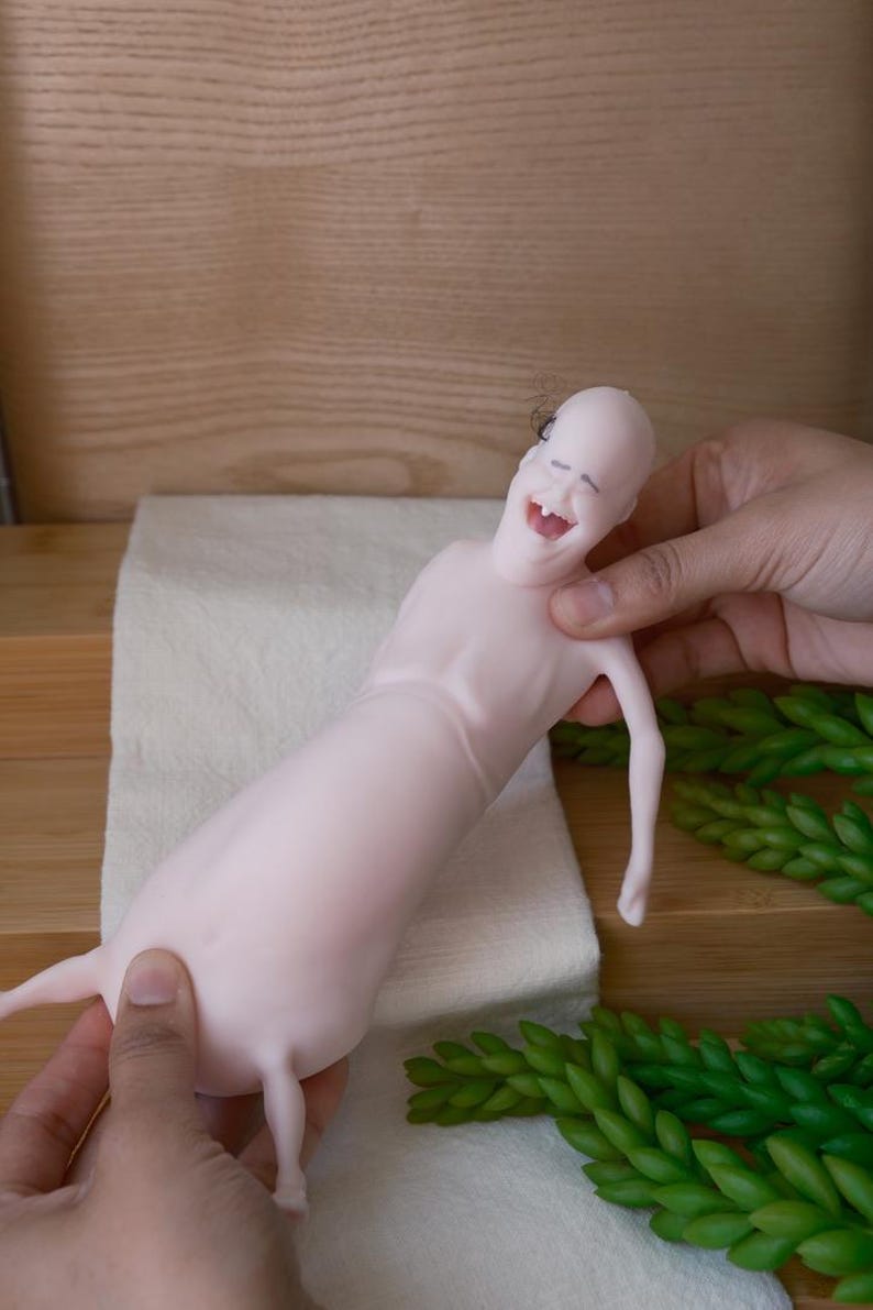 May include: A light pink, whimsical figurine with a smiling face and small limbs. The figure is held against a backdrop of a wooden surface, a white cloth, and green decorative plants. The figurine's design is cartoonish.